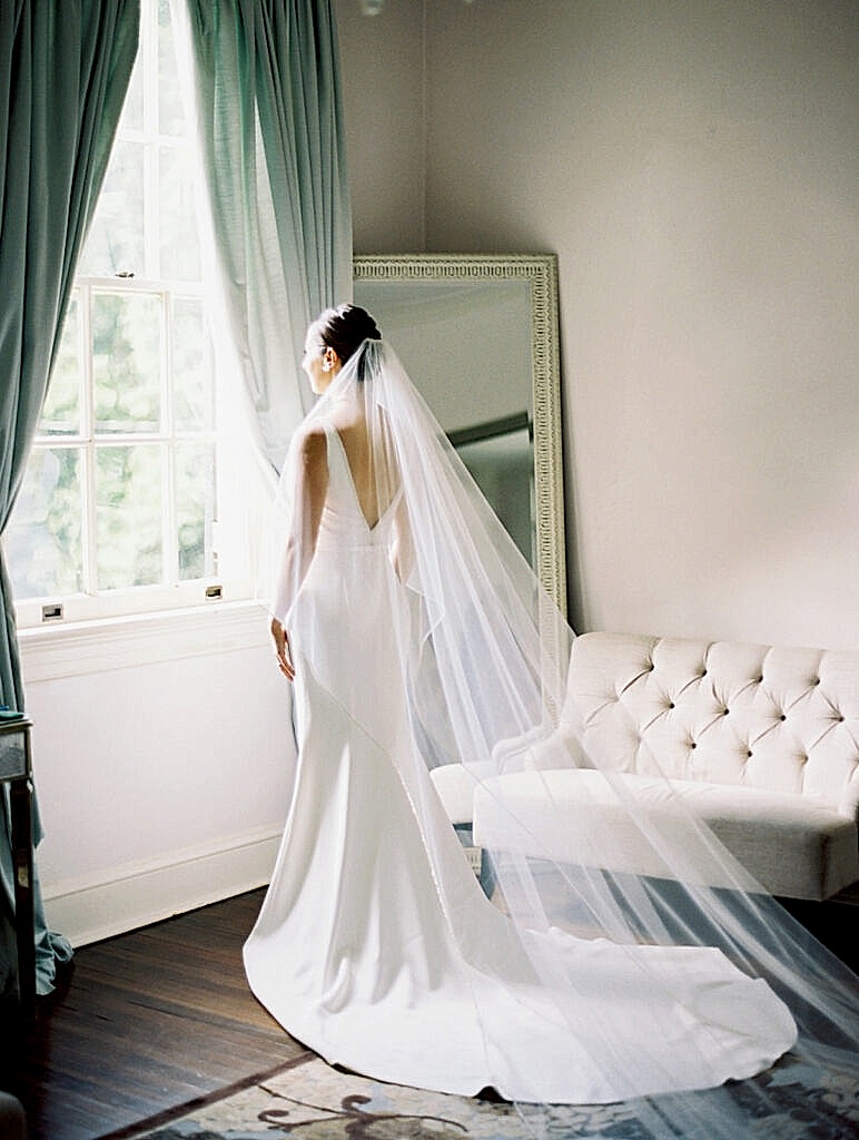 Bride standing by window brighly lit by the sun as her veil cascades behind her.